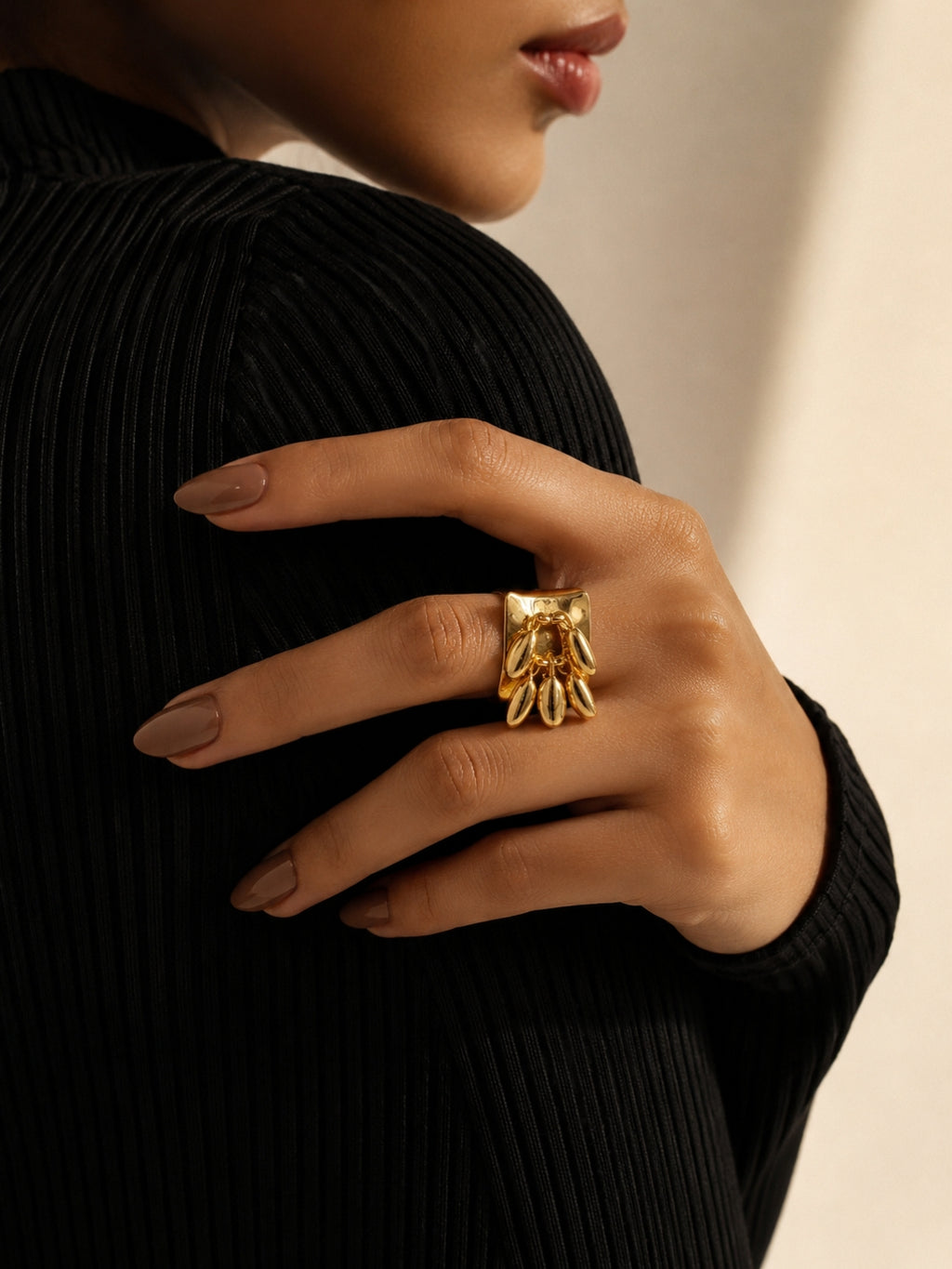A close-up of a model's hand wearing a chunky gold ring with a sculptural, textured design, styled with a black ribbed top against a warm beige background.