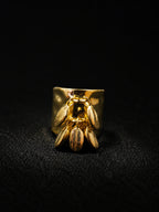 A gold ring featuring a sculpted bee design on a wide band, photographed against a dark textured background.