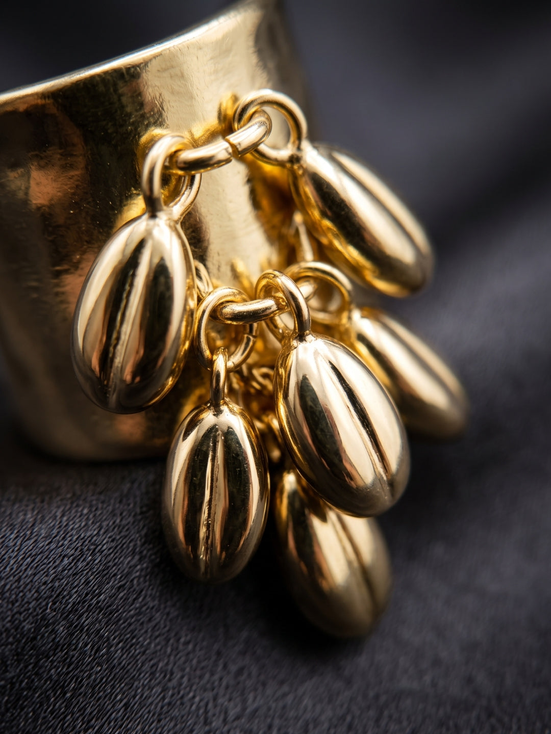 A close-up of a shiny gold jewelry piece with multiple dangling teardrop-shaped charms attached to a curved gold base, set against a dark textured fabric background.