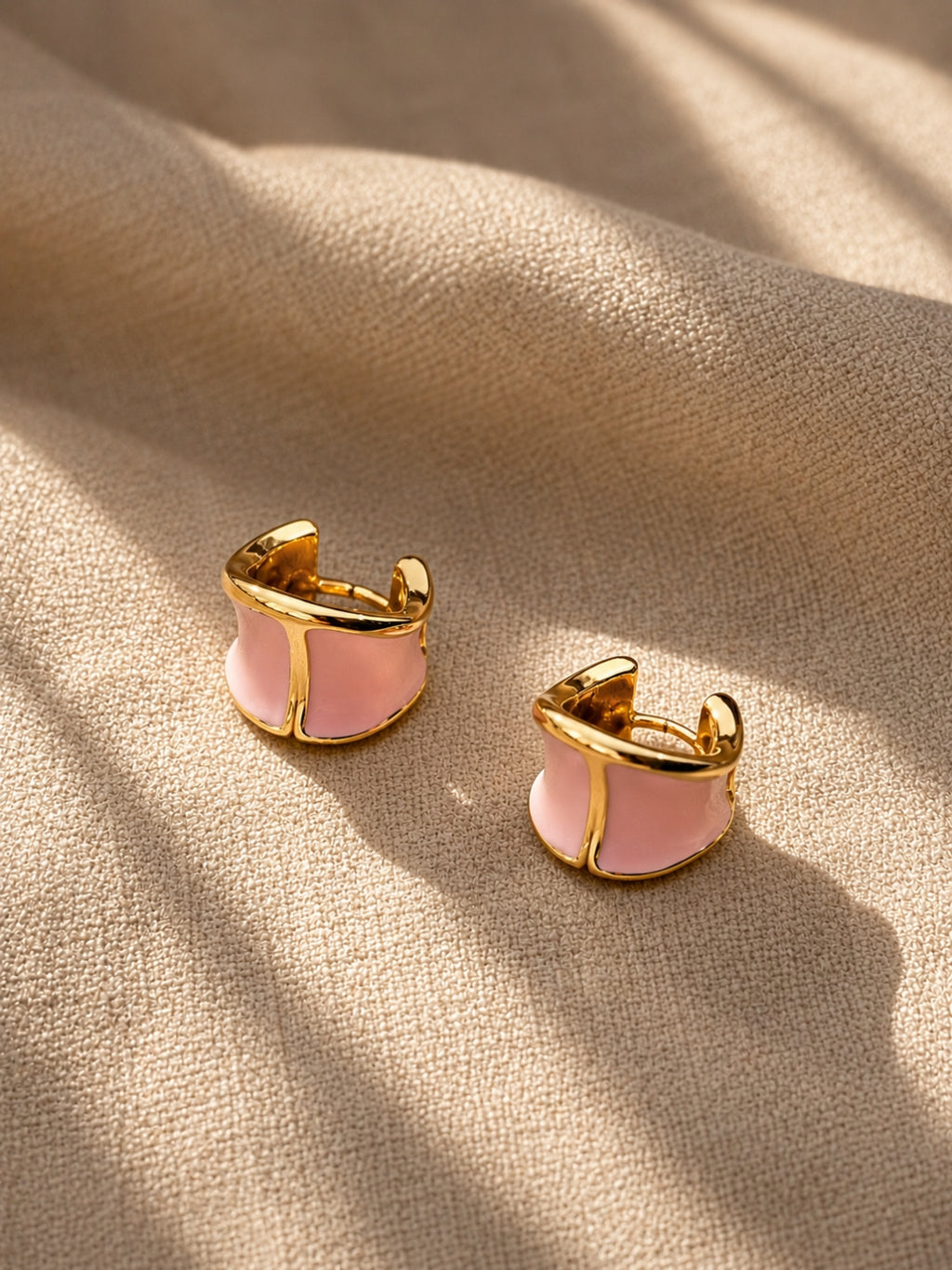 A pair of gold-toned huggie hoop earrings with a light pink enamel center, resting on a textured beige fabric background with soft shadows.