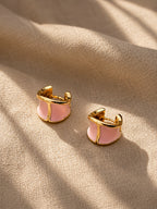 A pair of gold-toned huggie hoop earrings with a light pink enamel center, resting on a textured beige fabric background with soft shadows.