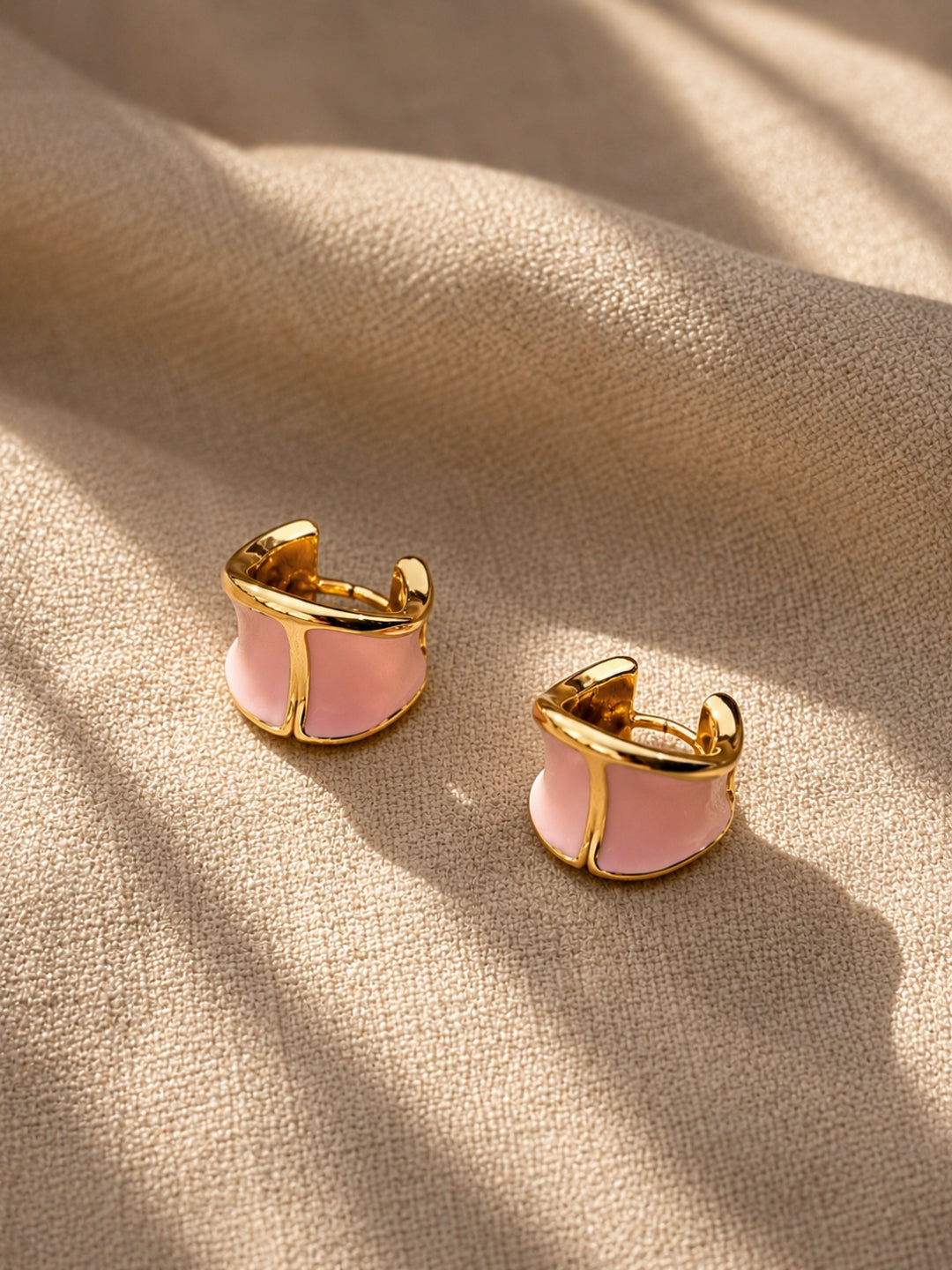 A pair of gold-toned huggie hoop earrings with a light pink enamel center, resting on a textured beige fabric background with soft shadows.
