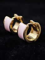 A pair of gold huggie hoop earrings with a curved white enamel inlay, displayed on a black textured background.