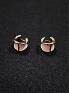 A pair of gold-toned hoop earrings with a central white and light pink geometric inlay, displayed on a textured black fabric background.