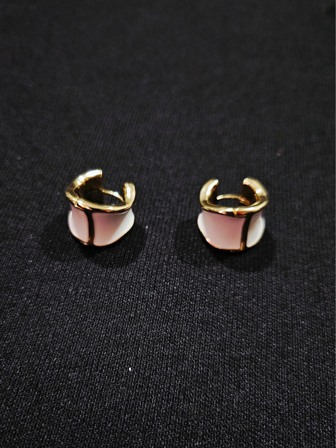 A pair of gold-toned hoop earrings with a central white and light pink geometric inlay, displayed on a textured black fabric background.