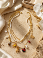 A layered gold and white pearl necklace with multiple dangling charms, including a red clover pendant, displayed on a textured beige stone surface with draped cream fabric and white orchid flowers.