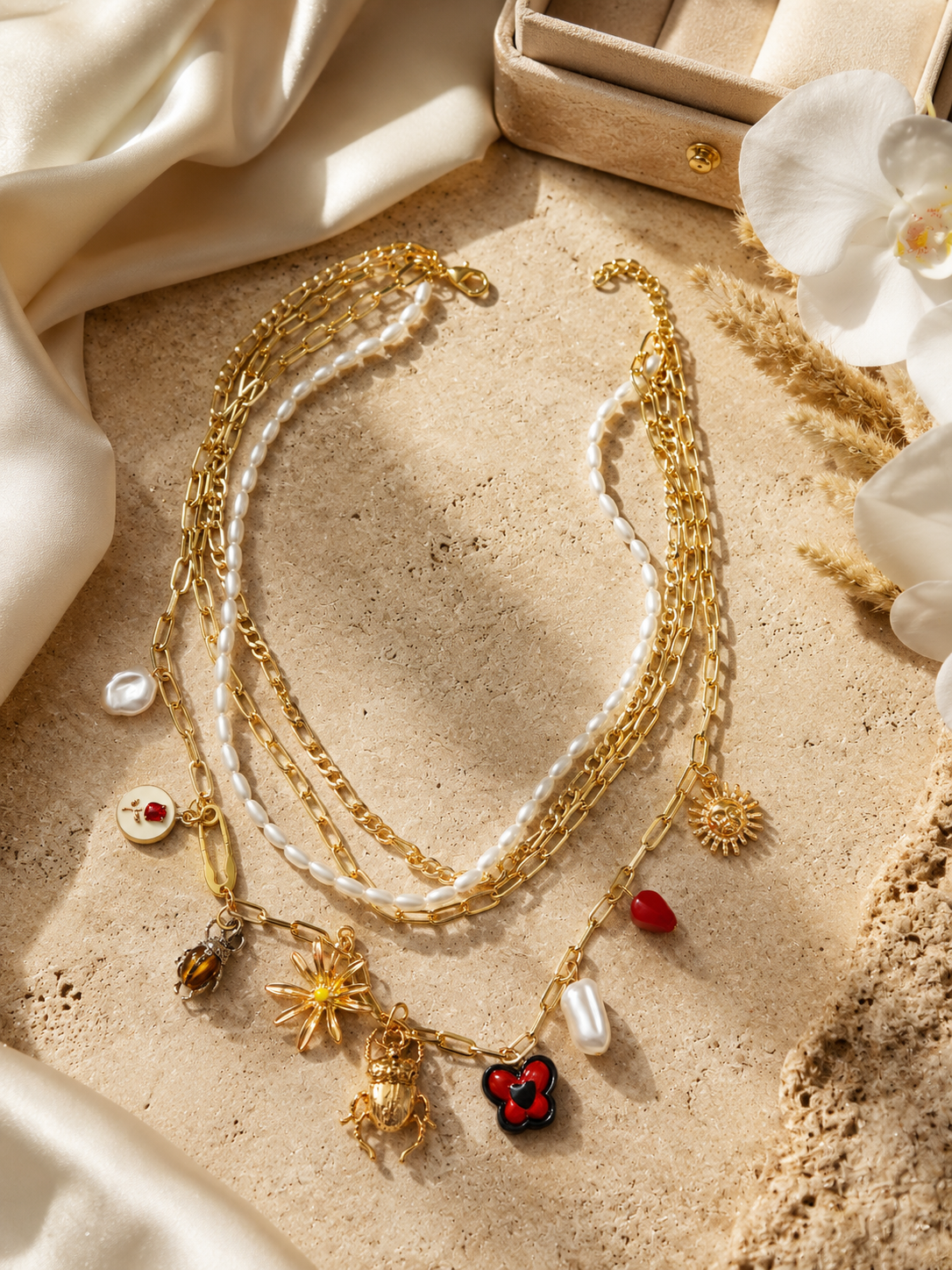 A layered gold and white pearl necklace with multiple dangling charms, including a red clover pendant, displayed on a textured beige stone surface with draped cream fabric and white orchid flowers.