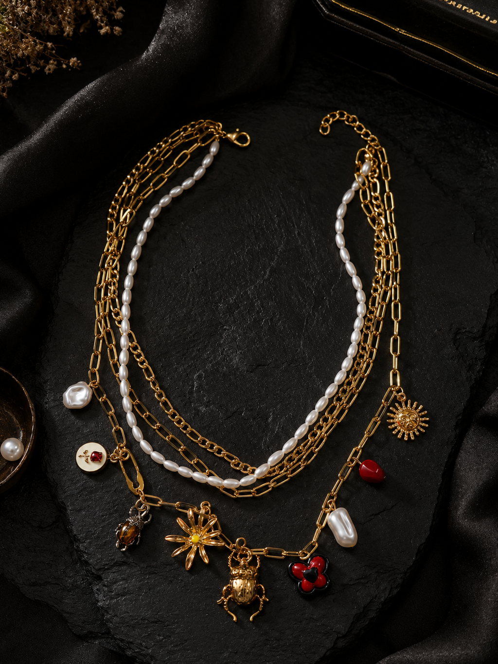 A multi-layered gold chain necklace with white pearl strands and various charms, including a sun, a spider, a heart, and a small red gem, displayed on a black textured surface.