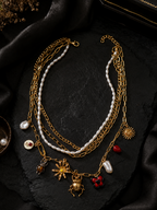 A multi-layered gold chain necklace with white pearl strands and various charms, including a sun, a spider, a heart, and a small red gem, displayed on a black textured surface.