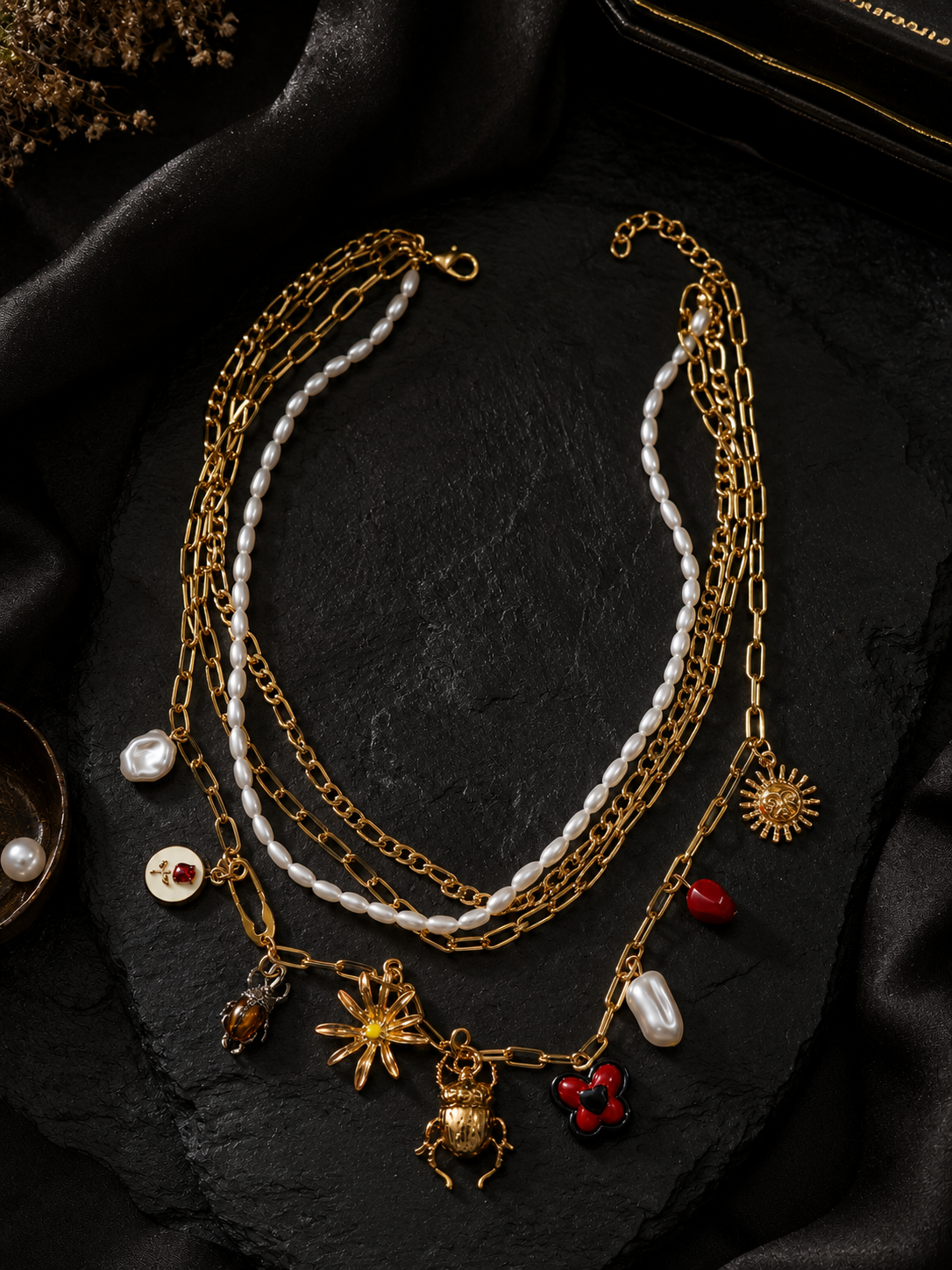 A multi-layered gold chain necklace with white pearl strands and various charms, including a sun, a spider, a heart, and a small red gem, displayed on a black textured surface.
