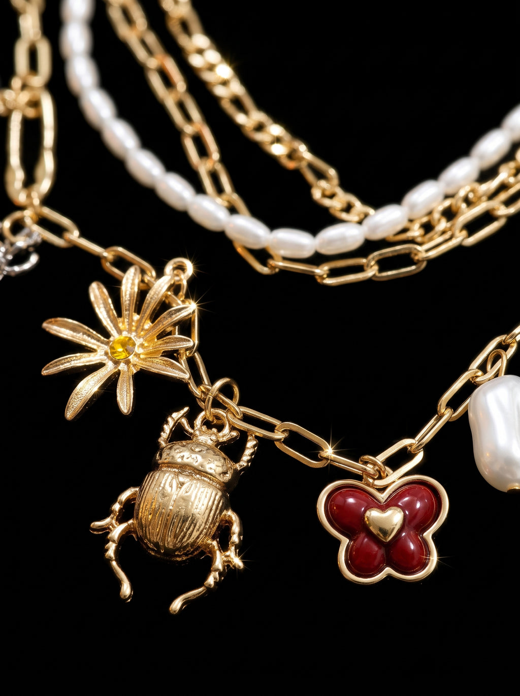 A close-up of a gold-tone layered necklace featuring a flower charm with a yellow center, a beetle pendant, a red clover charm with a gold heart, and white pearl-like beads on a black background.