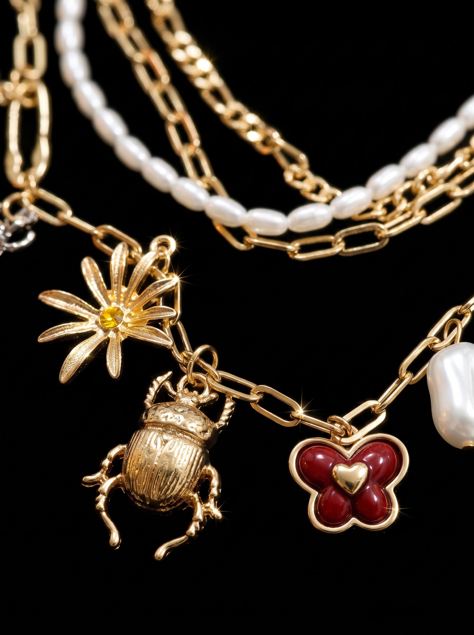 A close-up of a gold-tone layered necklace featuring a flower charm with a yellow center, a beetle pendant, a red clover charm with a gold heart, and white pearl-like beads on a black background.