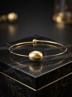 A gold-plated bangle bracelet with a textured spherical bead, displayed on a black marble block.