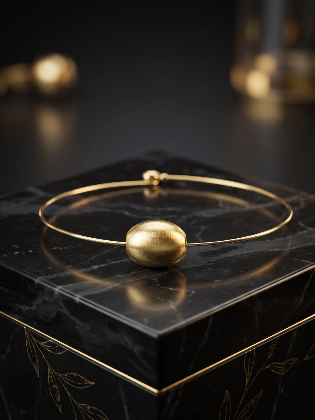 A gold-plated bangle bracelet with a textured spherical bead, displayed on a black marble block.