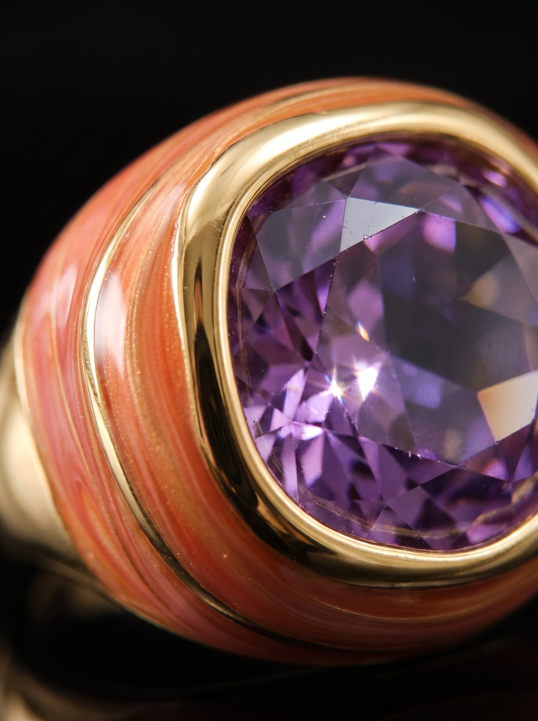 Close-up of a ring featuring a large faceted purple gemstone in a gold bezel setting, mounted on a cylindrical gold and pinkish-orange striped band against a black background.