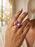 A close-up of a person's hand wearing a rose gold ring with a large, cushion-cut purple gemstone, set against a softly blurred warm indoor background.
