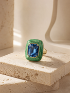 A gold ring with a textured green bezel holding a rectangular blue gemstone, displayed on a beige stone block.
