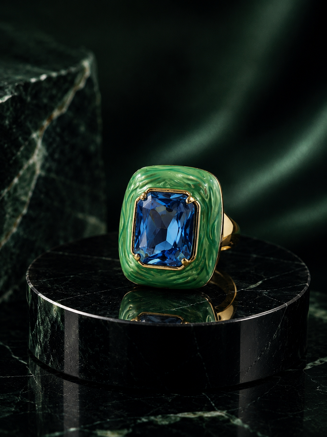 A gold ring with a textured green bezel and a large rectangular blue gemstone, displayed on a black marble pedestal against a dark green satin background.