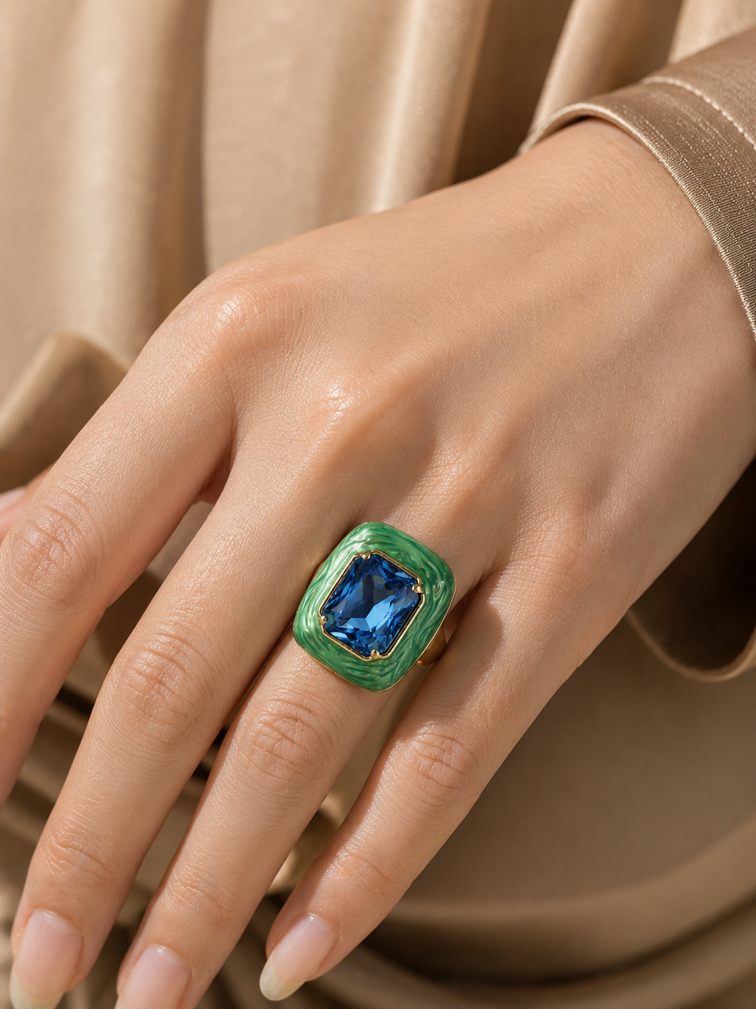A close-up of a hand wearing a gold ring with a square blue gemstone and a green enamel border, set against a soft beige satin background.