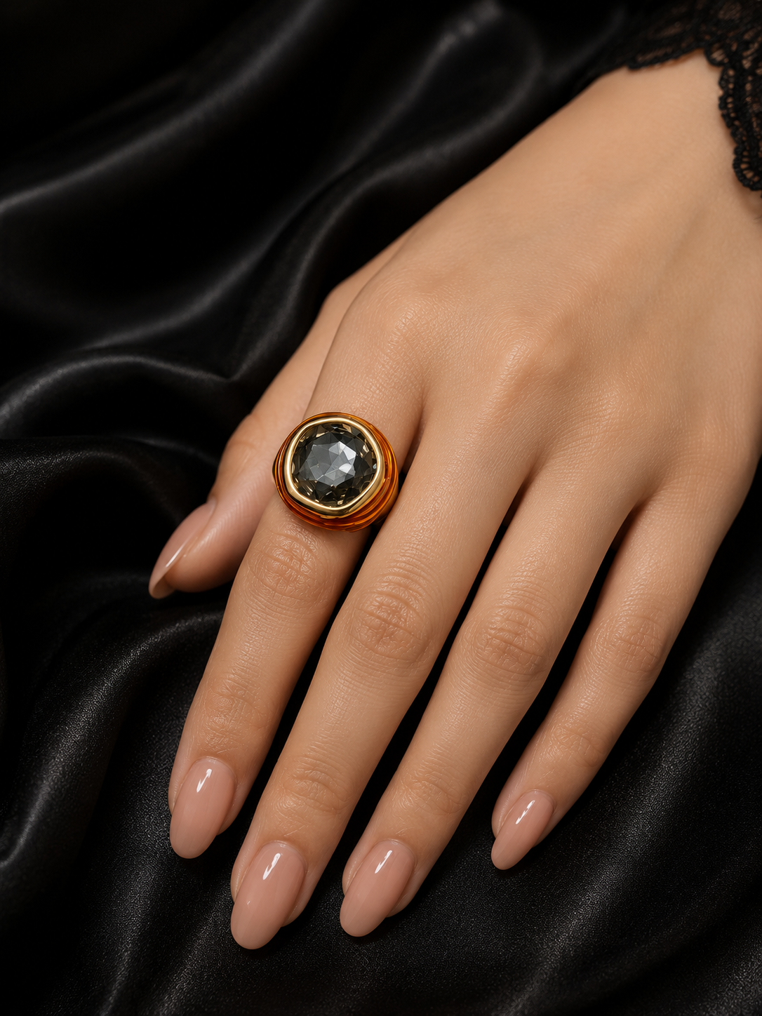 A gold signet-style ring with a large oval dark gray stone, worn on a model's hand against a draped black satin background.