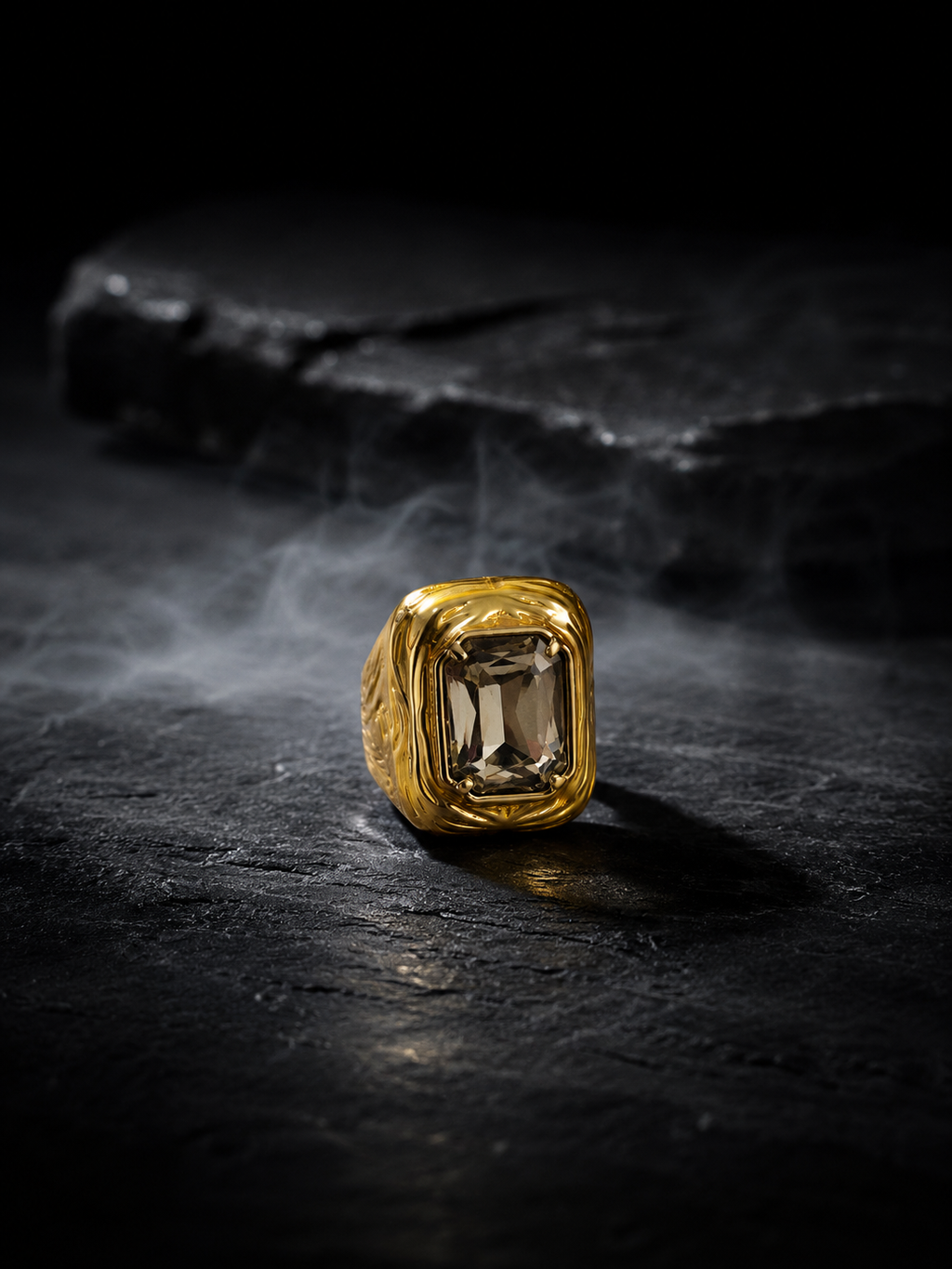 A gold signet-style ring with a rectangular smoky gemstone, photographed on a dark textured surface with smoke and a dark stone prop in the background.
