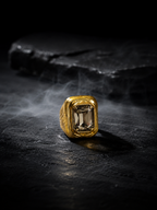 A gold signet-style ring with a rectangular smoky gemstone, photographed on a dark textured surface with smoke and a dark stone prop in the background.