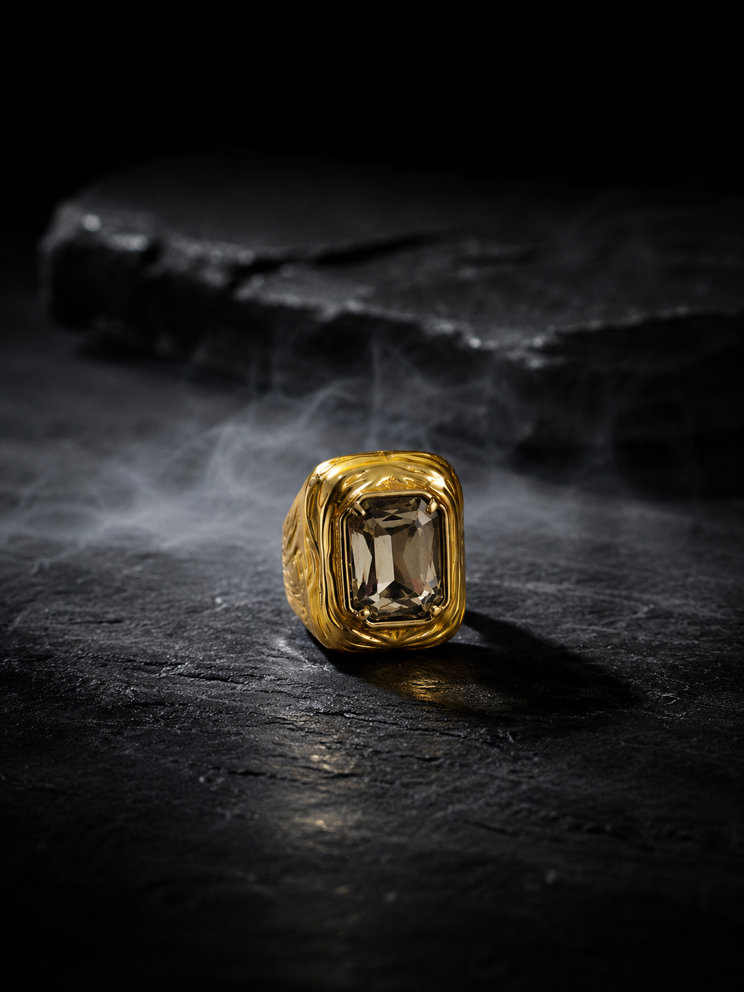 A gold signet-style ring with a rectangular smoky gemstone, photographed on a dark textured surface with smoke and a dark stone prop in the background.