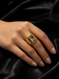 A gold signet-style ring with a large rectangular clear gemstone worn on a hand, set against a dark black satin fabric background.
