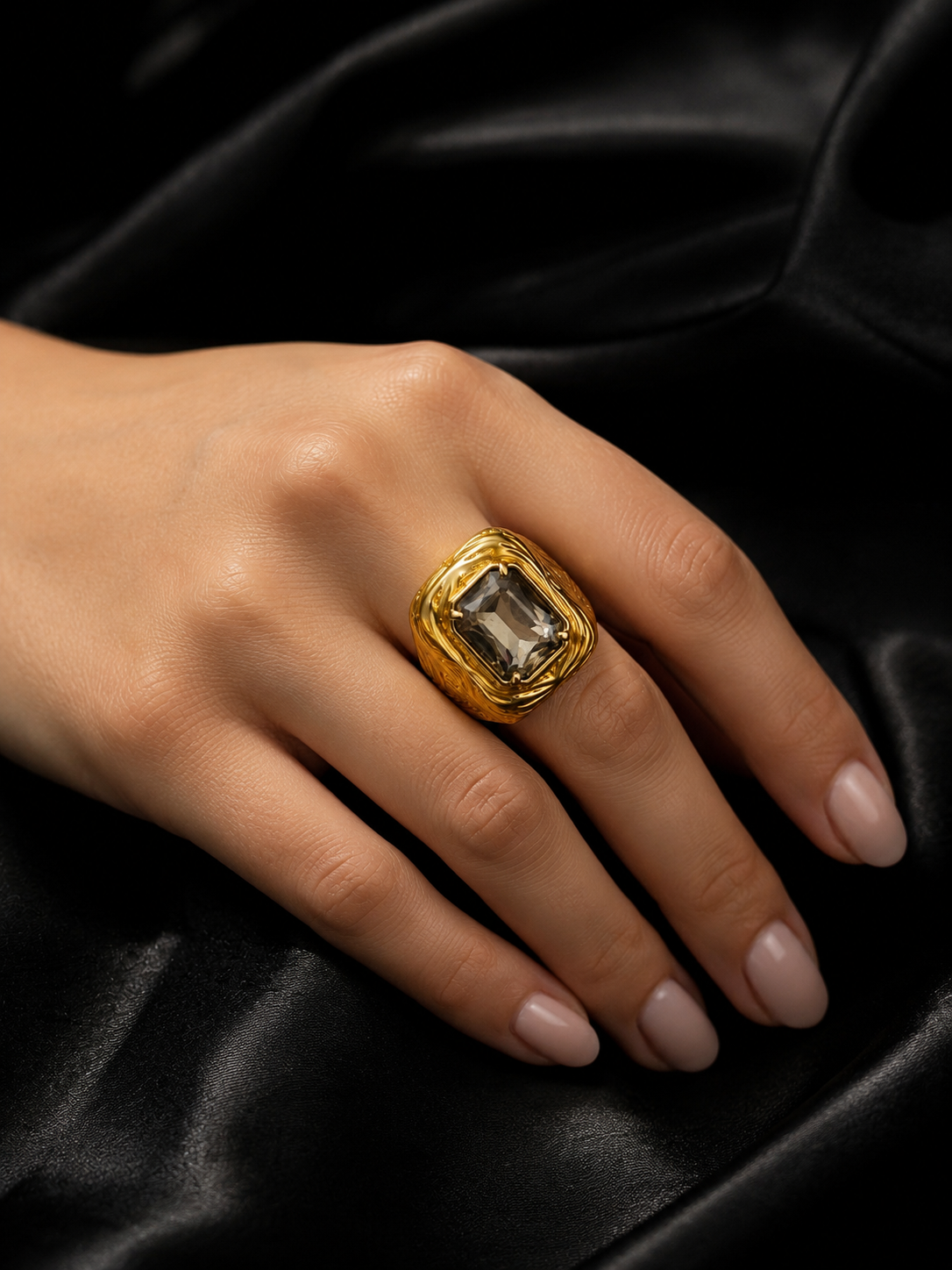 A gold signet-style ring with a large rectangular clear gemstone worn on a hand, set against a dark black satin fabric background.