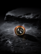 A round ring with a large faceted dark gray gemstone and a textured coppery-gold bezel, resting on a dark stone surface with dramatic smoke in the background.