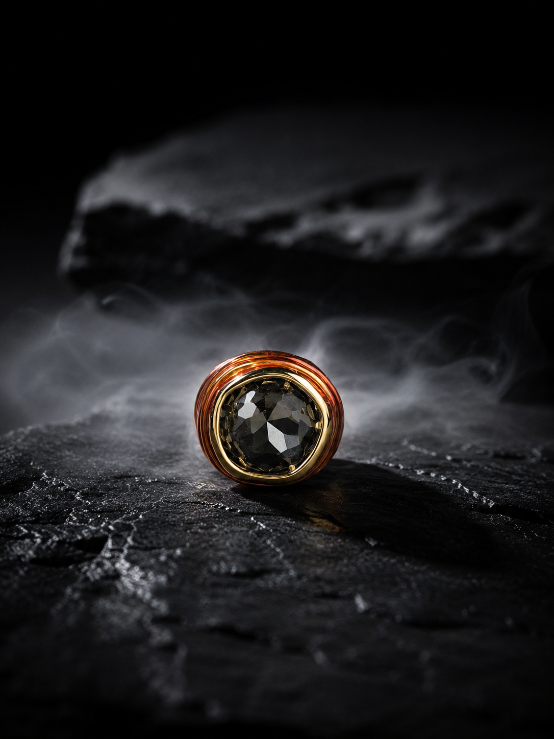 A round ring with a large faceted dark gray gemstone and a textured coppery-gold bezel, resting on a dark stone surface with dramatic smoke in the background.