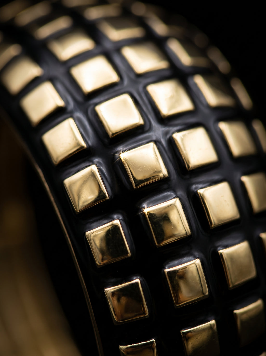 Close-up of a black studded leather belt showing a grid pattern of shiny gold square studs against a dark background.