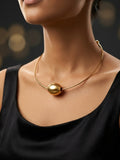 A close-up of a model wearing a delicate gold wire necklace with a large, polished gold sphere pendant at the center, styled with a black top against a dark, softly blurred background.