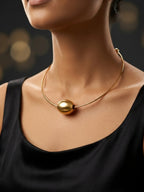A close-up of a model wearing a delicate gold wire necklace with a large, polished gold sphere pendant at the center, styled with a black top against a dark, softly blurred background.