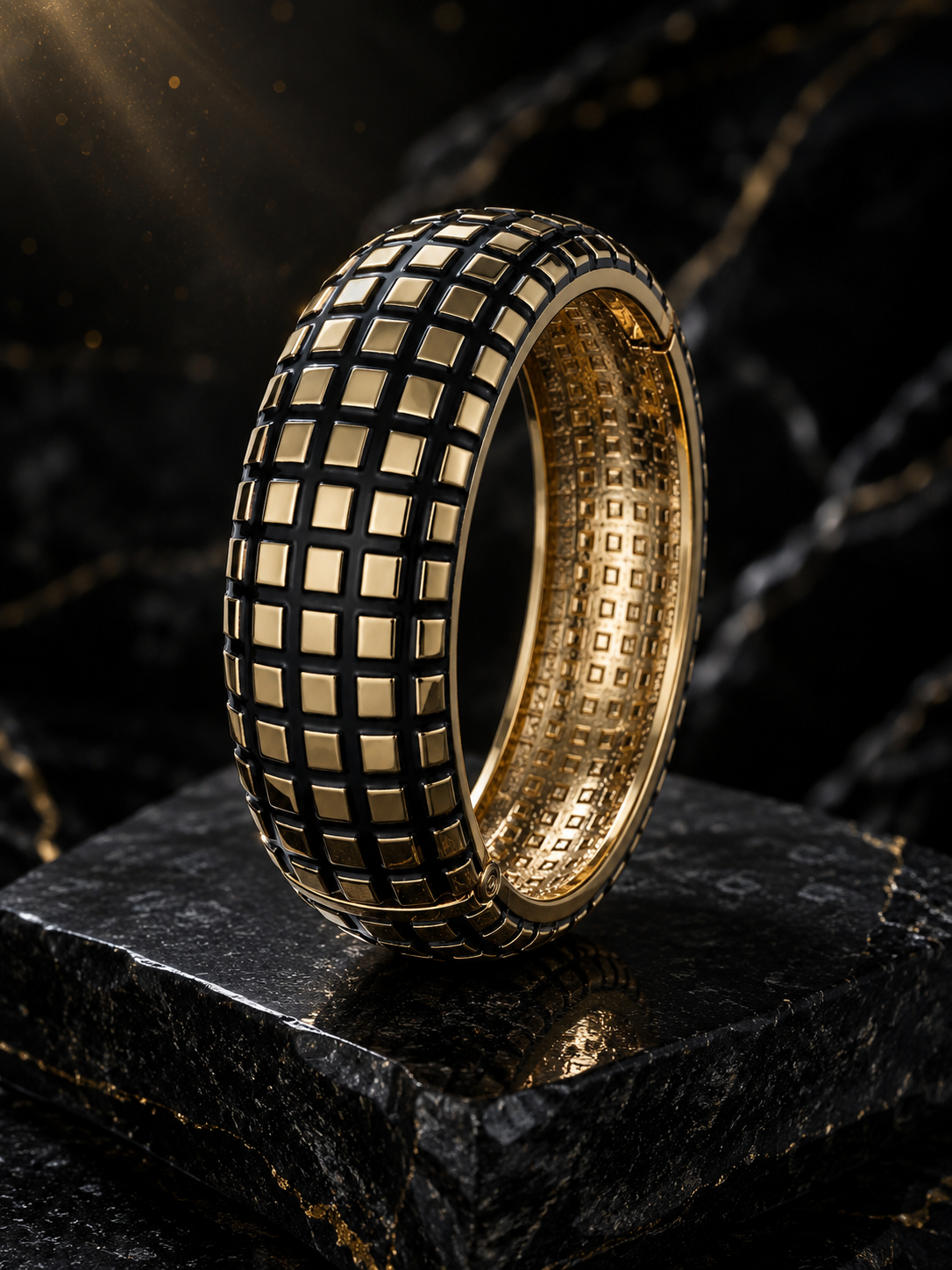 A wide gold ring with a black grid pattern on the exterior and a matching gold interior, displayed on a dark stone surface against a dark marble background.
