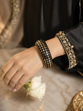 A close-up of a person's hand and wrist wearing two black beaded bangles with gold-toned geometric and floral patterns, styled with a white rose on a marble surface.