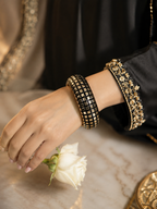 A close-up of a person's hand and wrist wearing two black beaded bangles with gold-toned geometric and floral patterns, styled with a white rose on a marble surface.