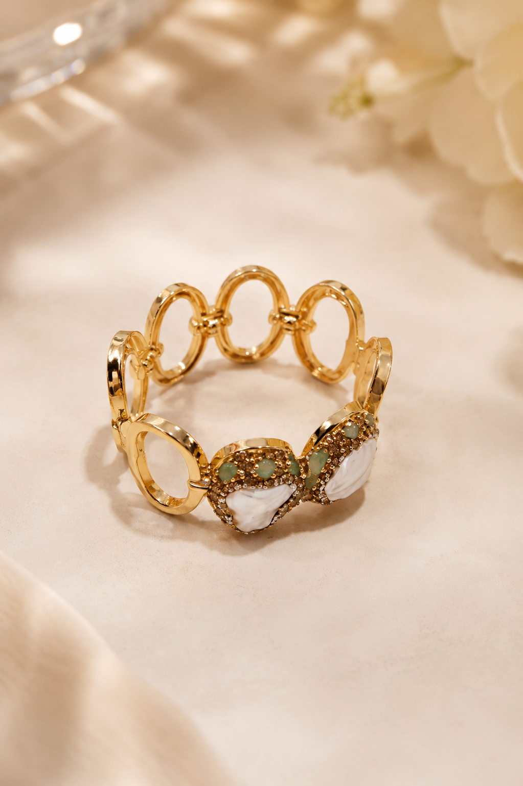 A gold-plated stretch ring featuring a series of interlocking oval links and a bow-shaped centerpiece adorned with white mother-of-pearl inlays and small green gemstones, resting on a soft beige fabric background.