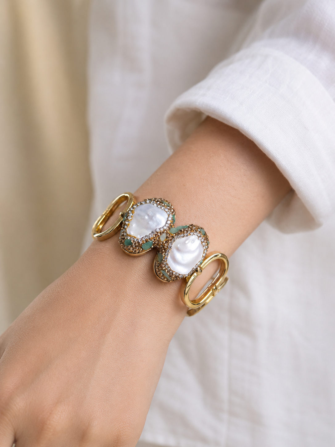 A close-up of a gold-tone cuff bracelet worn on a person's wrist, featuring two large white mother-of-pearl heart-shaped cabochons surrounded by textured gold detailing and small green accents, with a white shirt cuff visible.