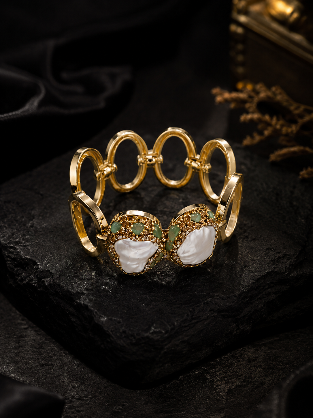 A gold-plated bangle bracelet with interlocking circular links and two large oval sections featuring white mother-of-pearl shell inlays and small green stones, displayed on a dark textured surface.