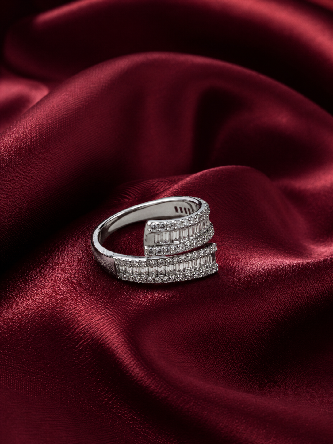 A silver wrap-style ring with pavé-set clear stones and baguette-cut stones, displayed on draped deep red satin fabric.