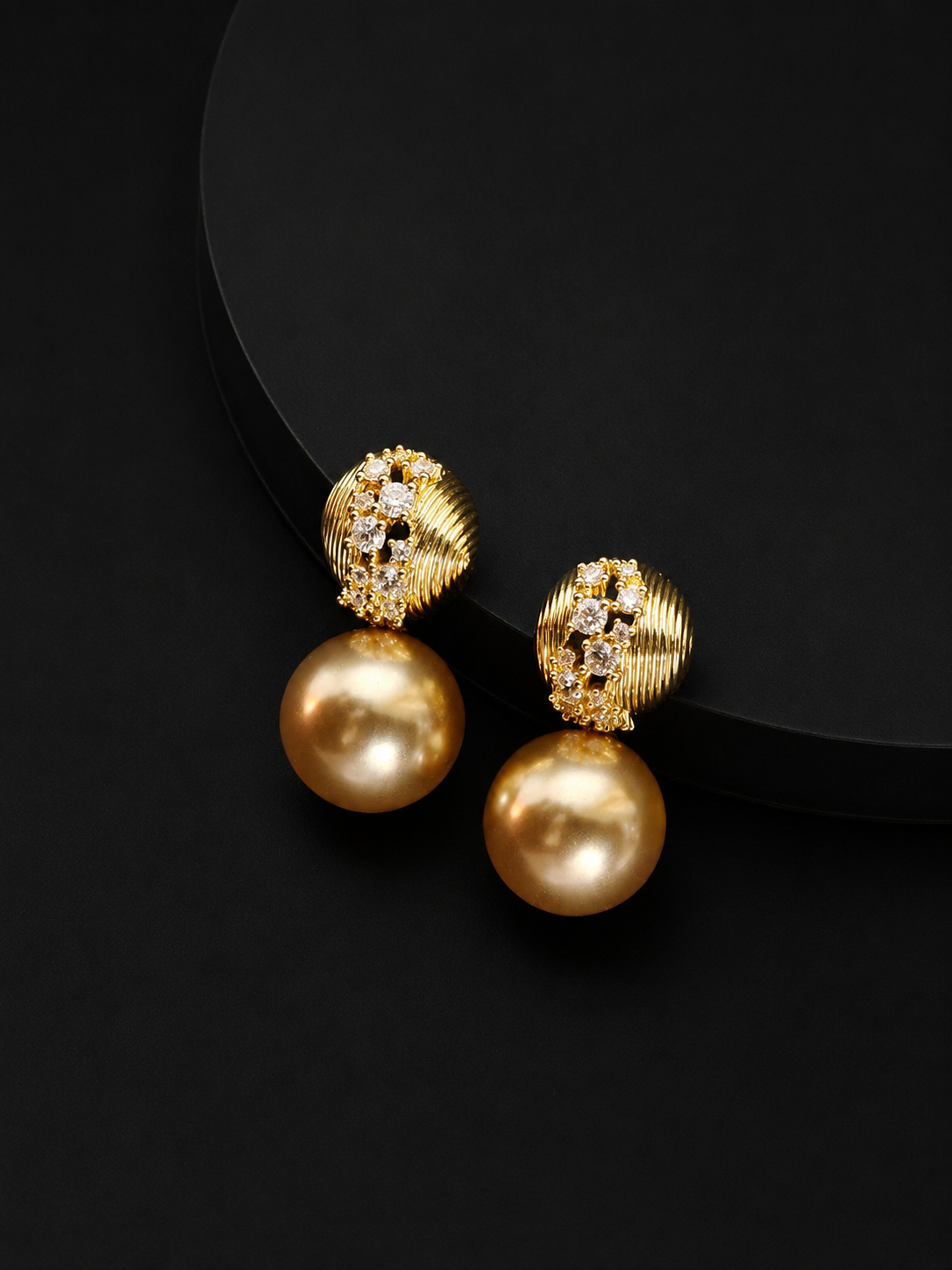 A pair of gold drop earrings with textured, diamond-like studs and large round gold pearls, displayed on a black background.