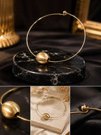 A gold-toned minimalist wire hoop earring with a matte spherical bead accent, displayed on a round black marble stand with a dark draped fabric background. Three smaller inset images at the bottom show close-up details of the earring from different angles.