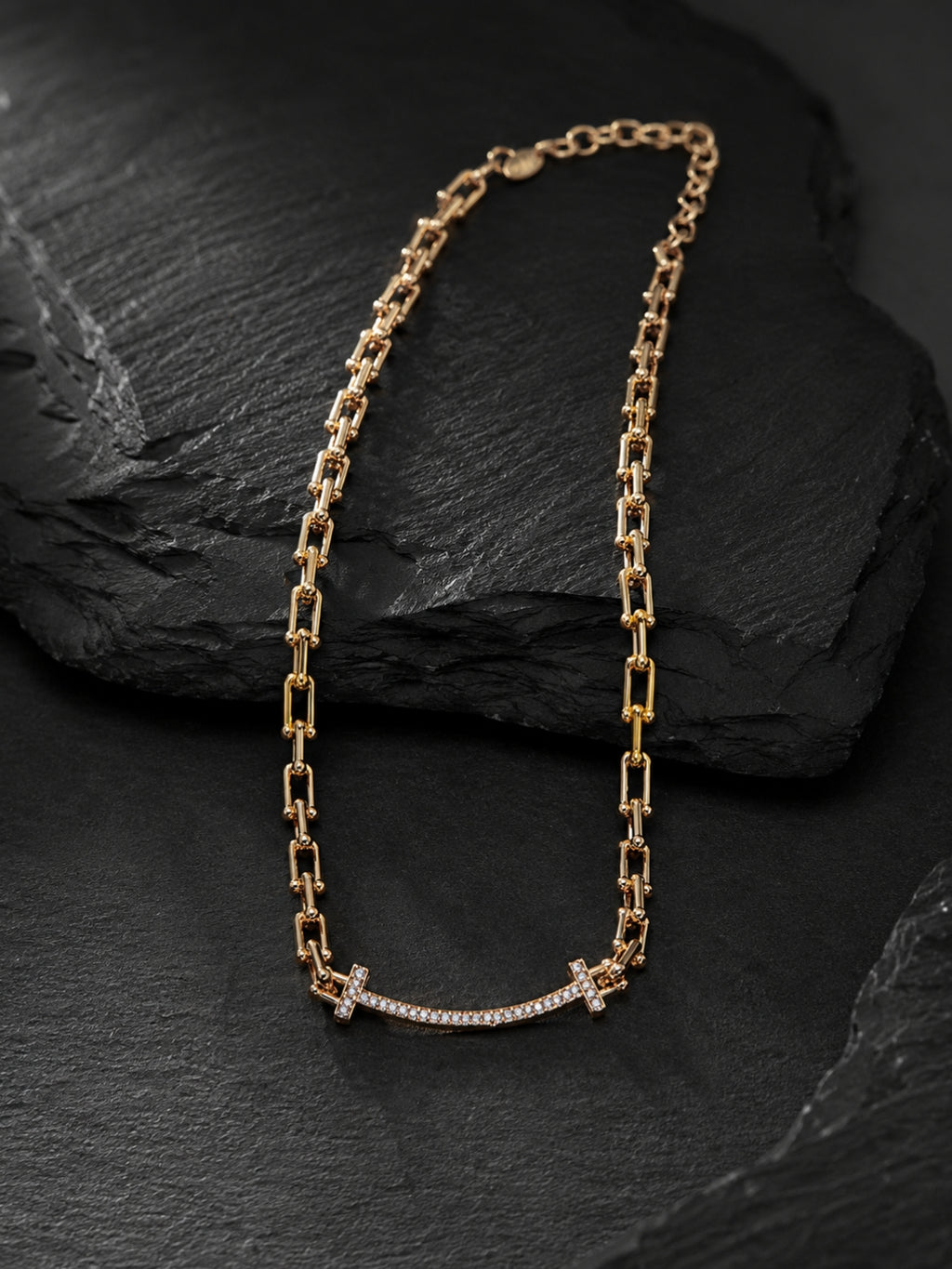 A gold chain necklace with a curved bar pendant set with small clear stones and a small cross at the center, displayed on a dark textured slate surface.