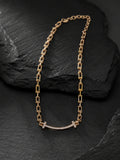 A gold chain necklace with a curved bar pendant set with small clear stones and a small cross at the center, displayed on a dark textured slate surface.