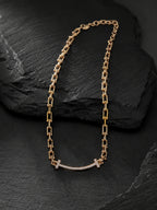 A gold chain necklace with a curved bar pendant set with small clear stones and a small cross at the center, displayed on a dark textured slate surface.
