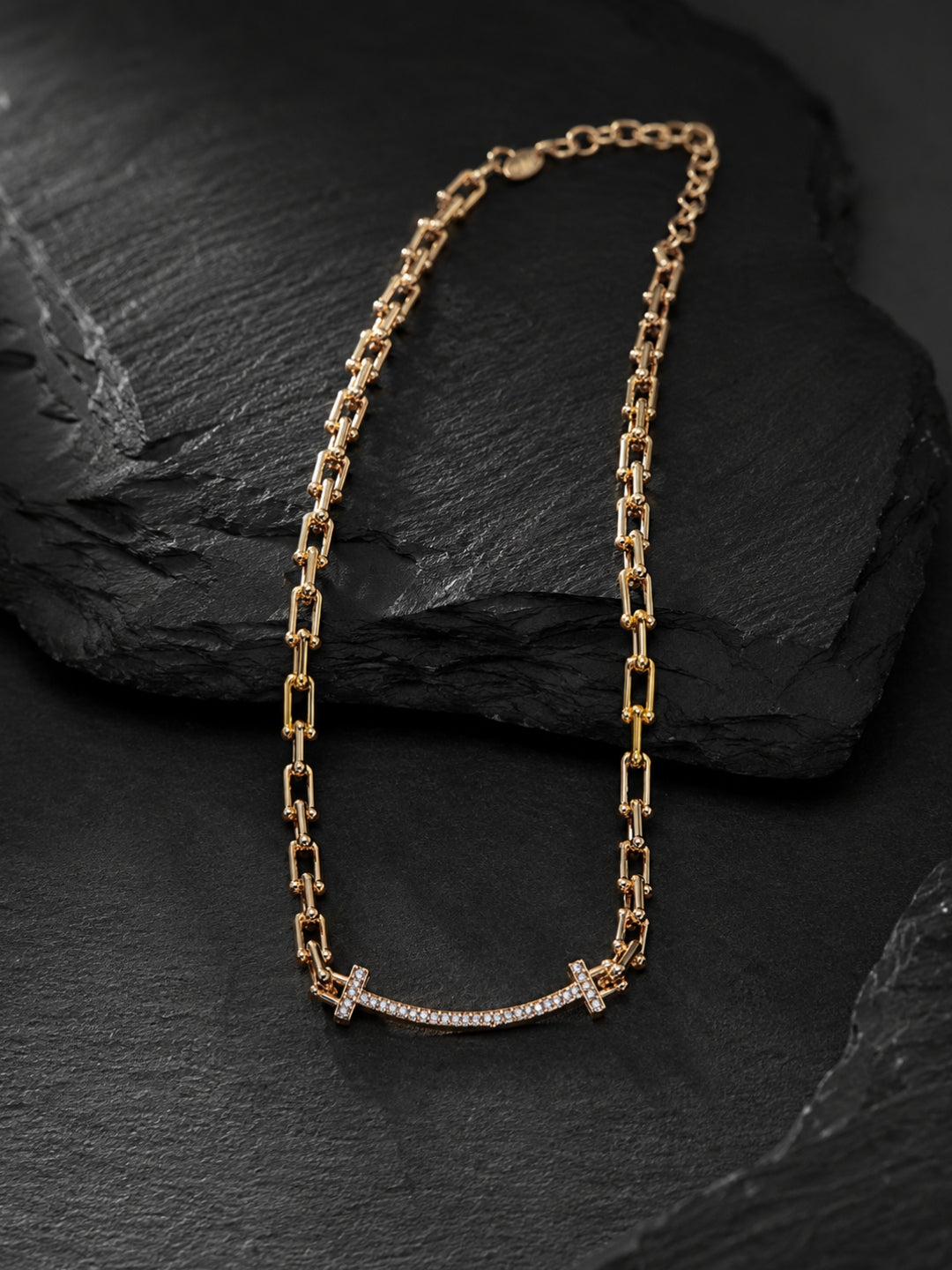 A gold chain necklace with a curved bar pendant set with small clear stones and a small cross at the center, displayed on a dark textured slate surface.