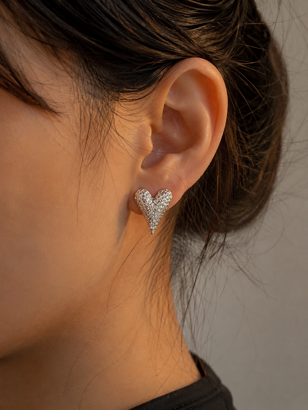 A close-up of a person's ear wearing a small heart-shaped stud earring covered in sparkling clear stones on a plain neutral background.