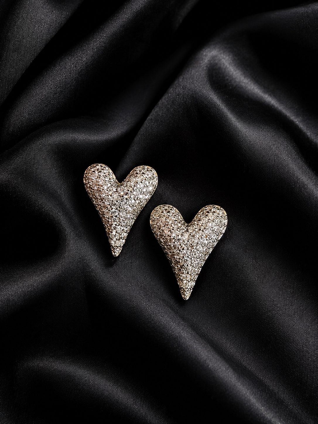 Two heart-shaped brooches covered in small clear stones, displayed on a draped black satin fabric background.