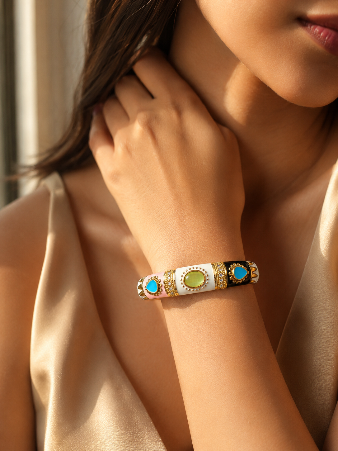 A close-up lifestyle shot of a woman wearing a gold-plated bangle bracelet with white enamel, a green stone, and turquoise blue stones.